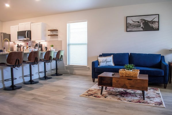A modern living room with a blue sofa, a coffee table, and a kitchen area with white cabinets and a bar stool setup.