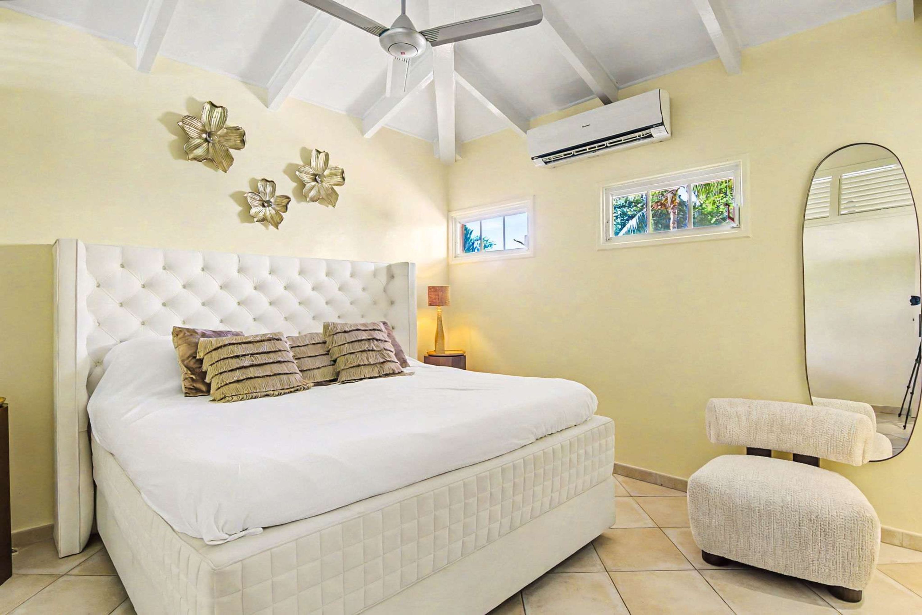 Un dormitorio con cabecera capitonada, cama con sábanas blancas, lámpara de noche, una silla pequeña, unidad de aire acondicionado y ventana con vistas.