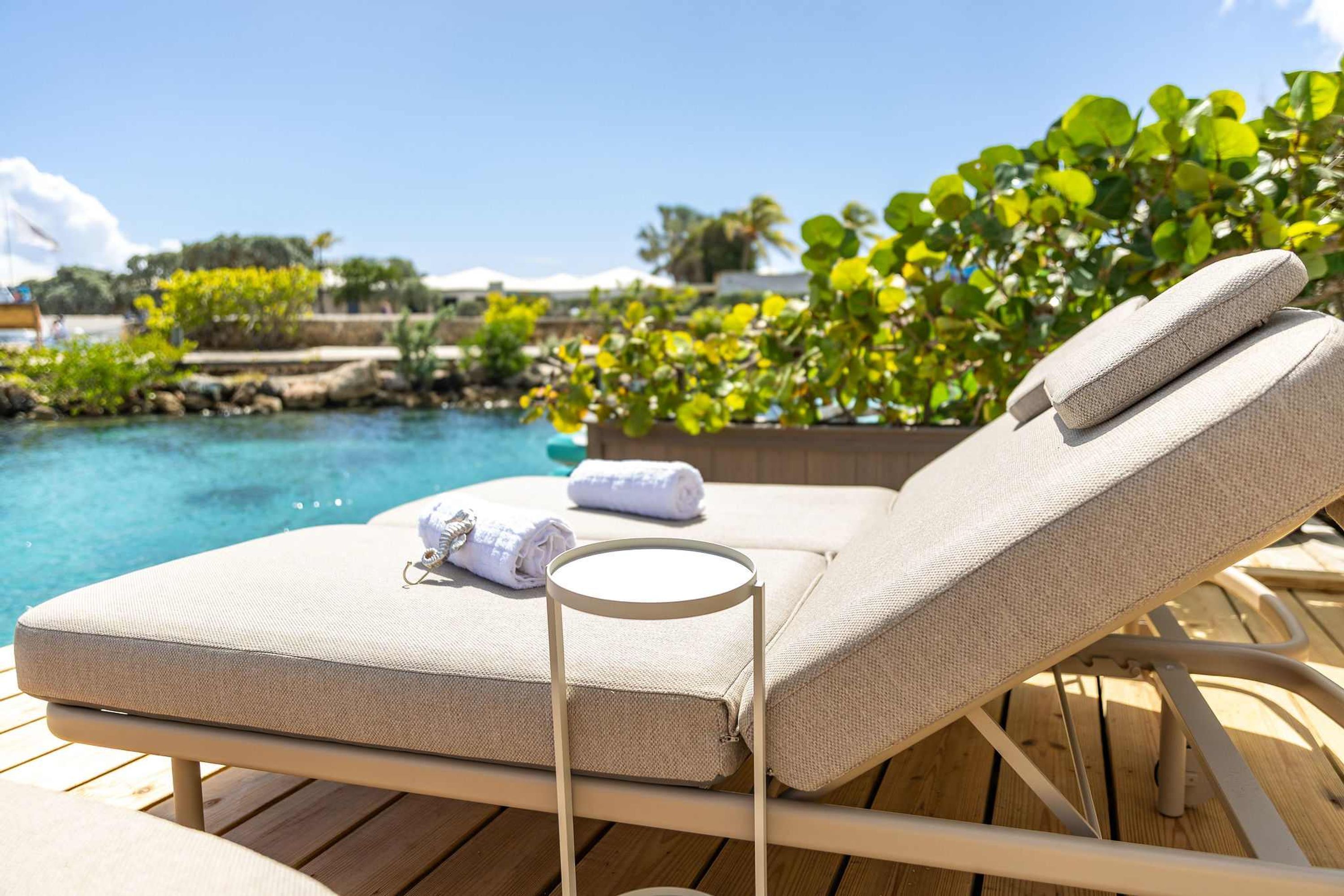 A sun lounger with a cushion and towel, near a pool with a wooden deck and greenery.