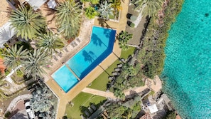 Luftaufnahme eines Resorts mit einem rechteckigen Pool, umgeben von Palmen und einem klaren, blauen Meer.