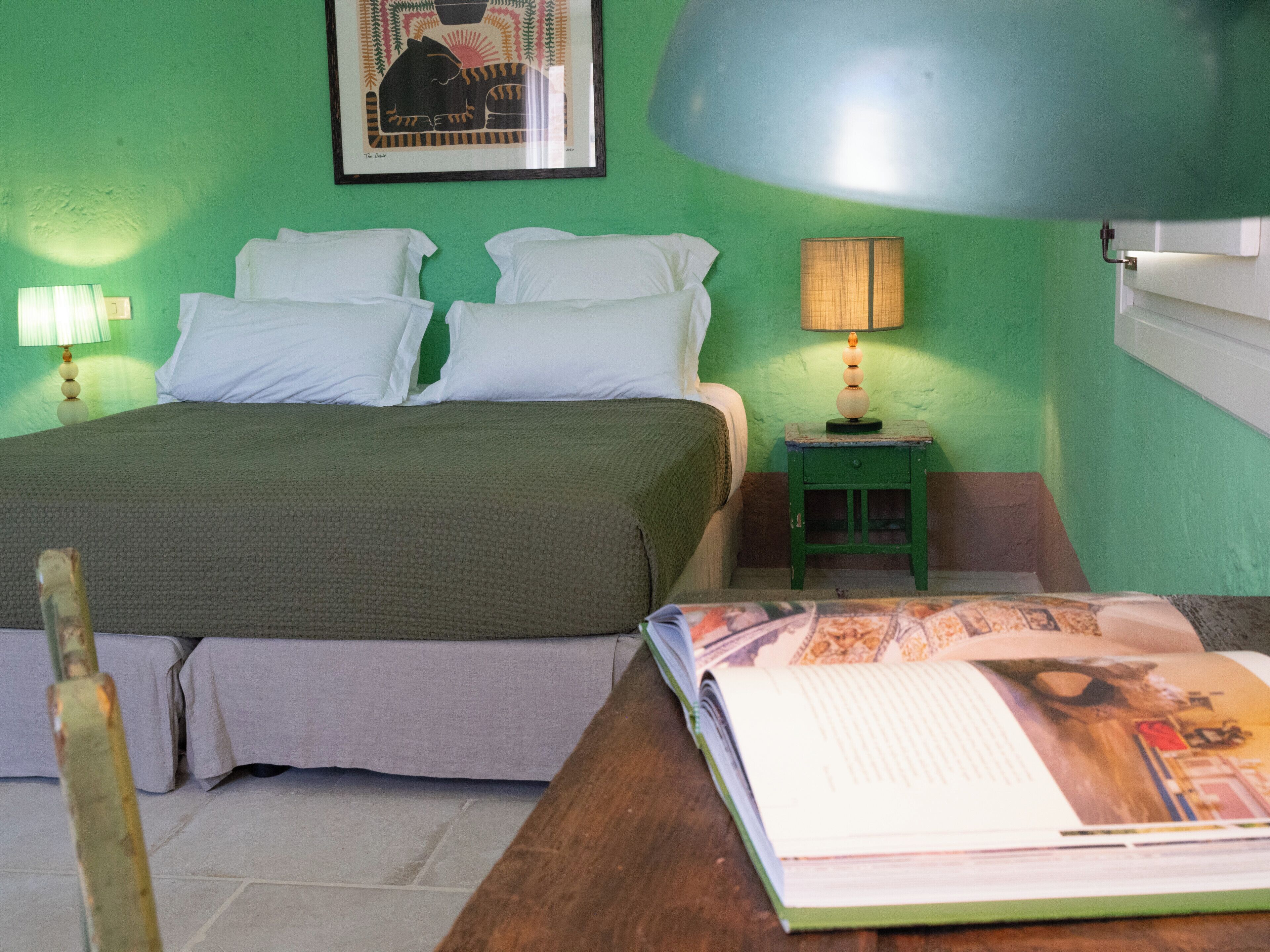 Una stanza con un letto, un comodino con una lampada, un quadro appeso al muro e un libro su un tavolo.