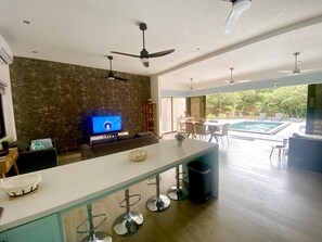 A modern kitchen with a TV, bar stools, and a pool area visible through the glass doors. - Cozy house, between beach and Nature (Brasilito)