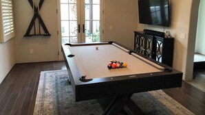 Une table de billard avec des billes, un téléviseur et un élément décoratif mural dans une pièce dotée d’un plancher en bois et d’une fenêtre.