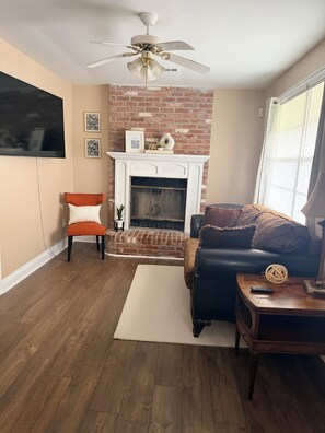 A living room with a brick fireplace, a ceiling fan, a television, a sofa, a chair, and a coffee table.
