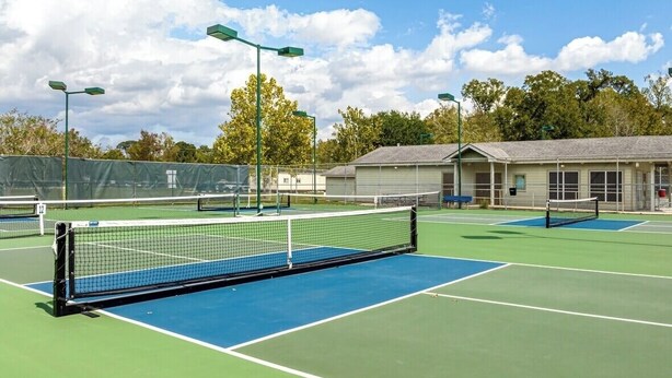 Camp de tennis amb una xarxa, envoltat d'arbres i un edifici amb teulada inclinada.