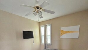 Un dormitori amb un llit, un ventilador de sostre, una televisió, una finestra amb cortines i un quadre a la paret.