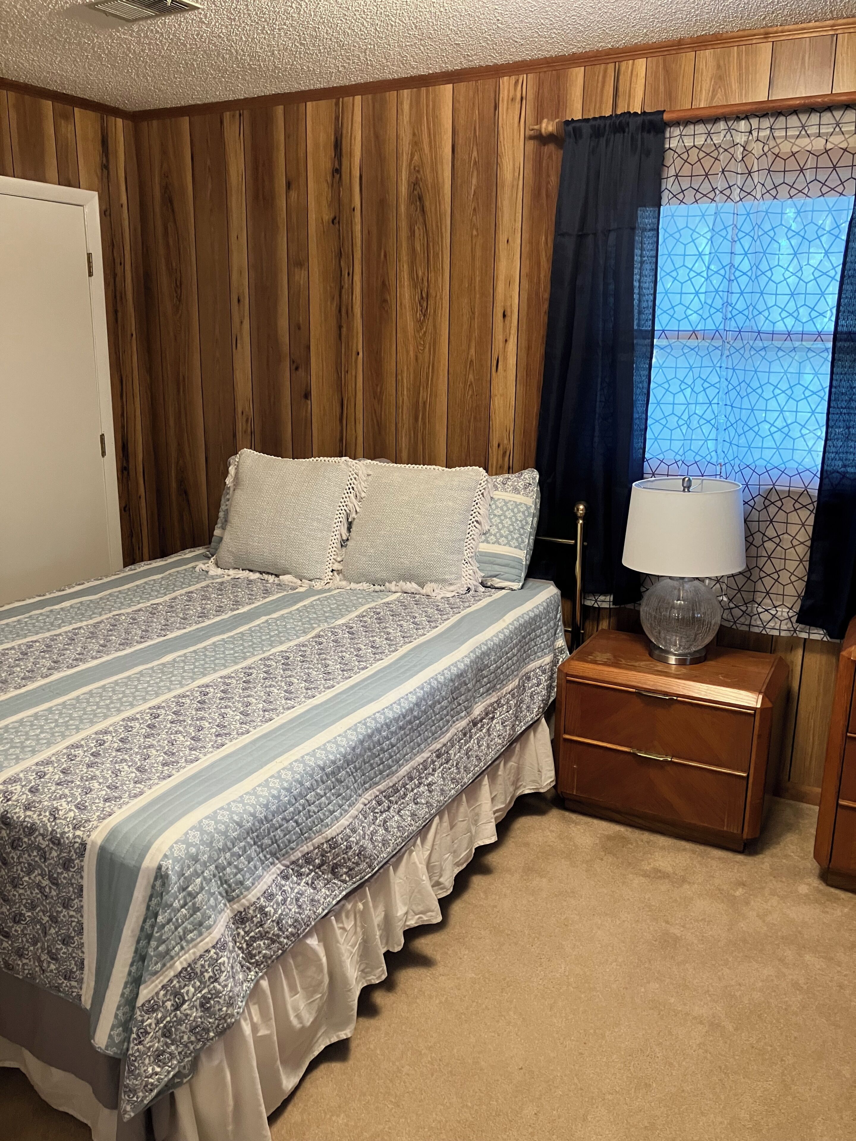 A bedroom with a wooden paneled wall, a bed with a patterned bedspread, a bedside table with a lamp, and a window with a geometric curtain.