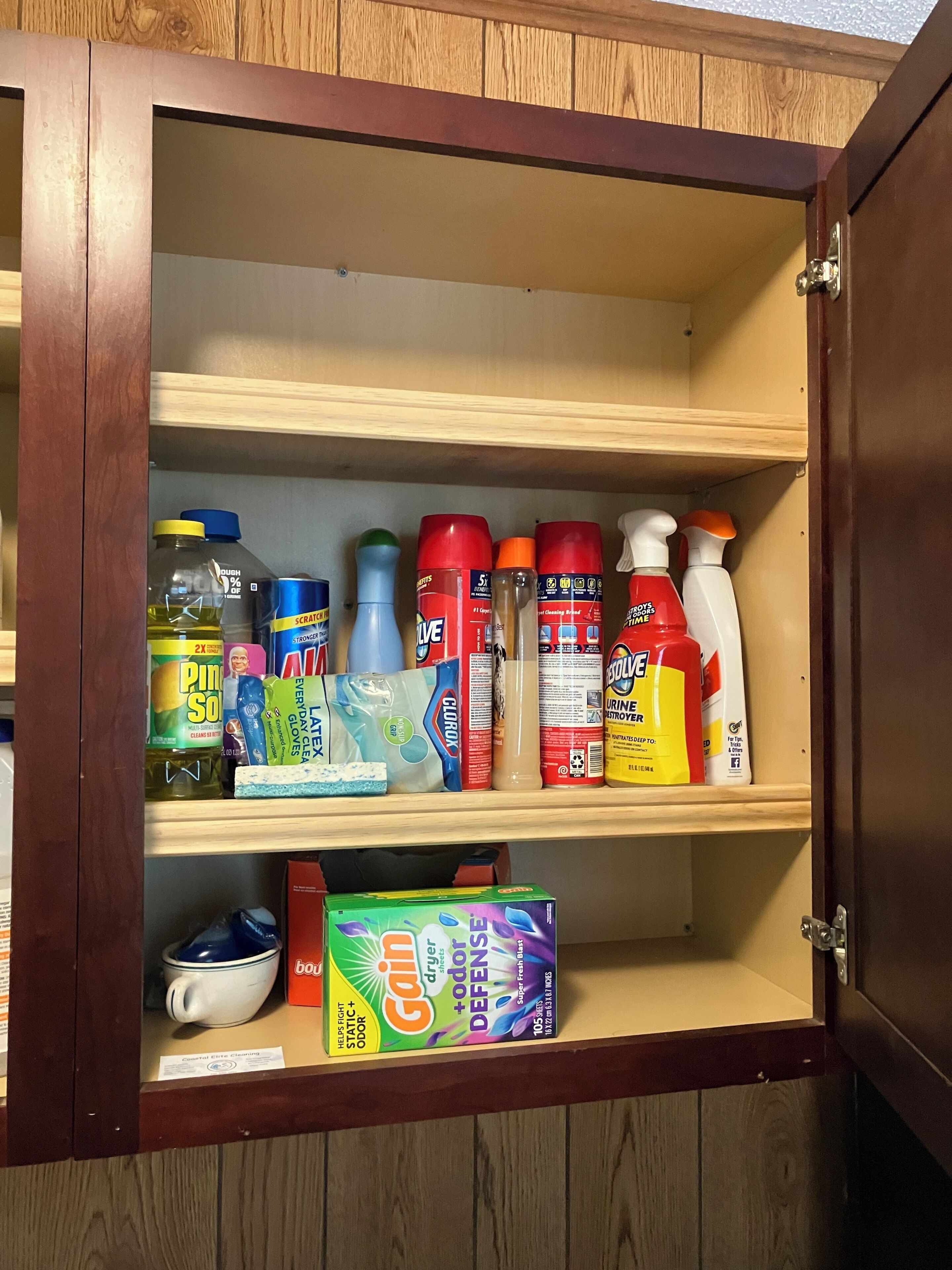 Un mueble de madera con repisas que contiene productos de limpieza, una caja de GAI® deodorant, y un tazón blanco.