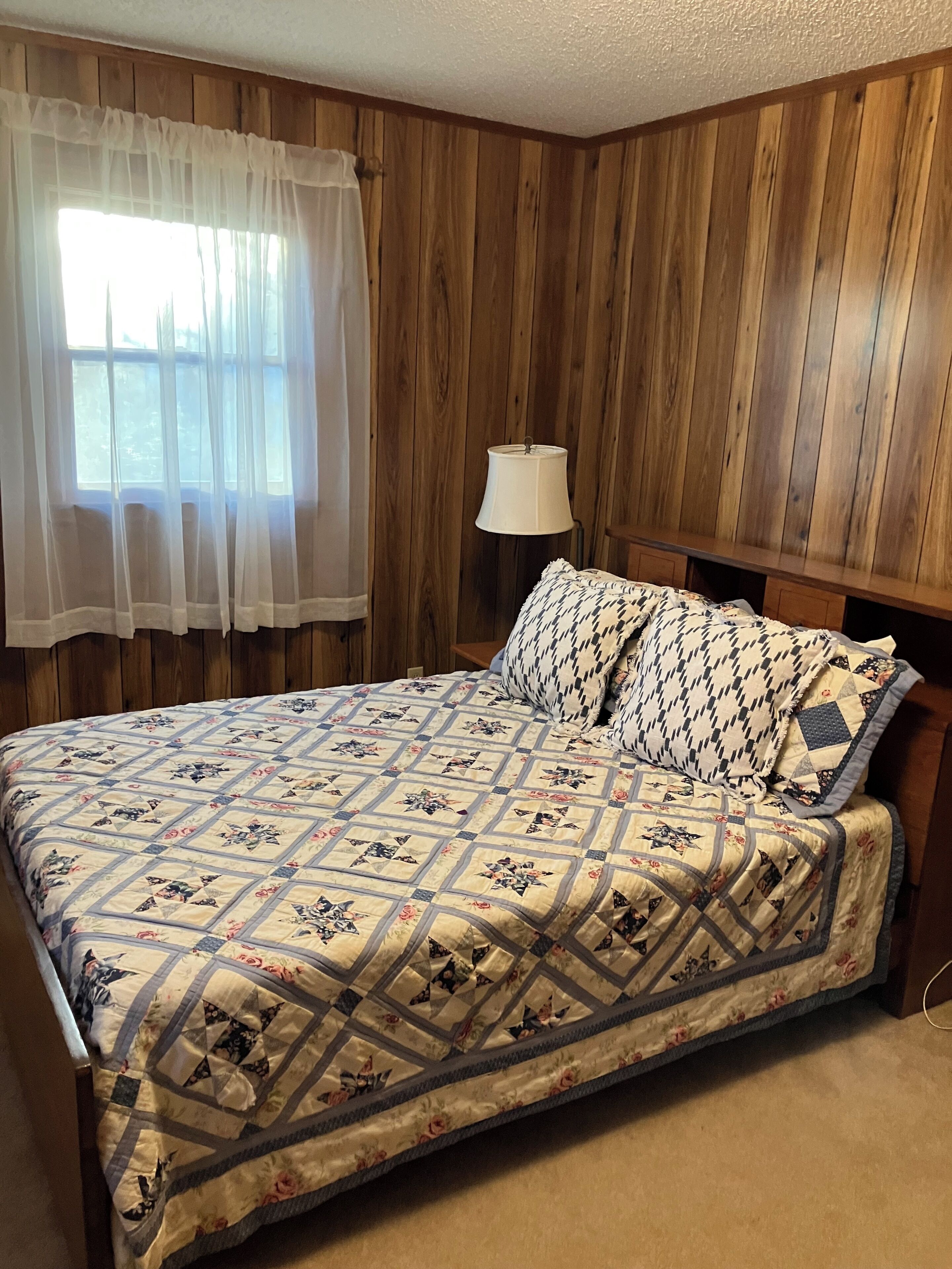 A bedroom with a wooden paneled wall, a bed with a floral quilt, a bedside lamp, and a window with sheer curtains.