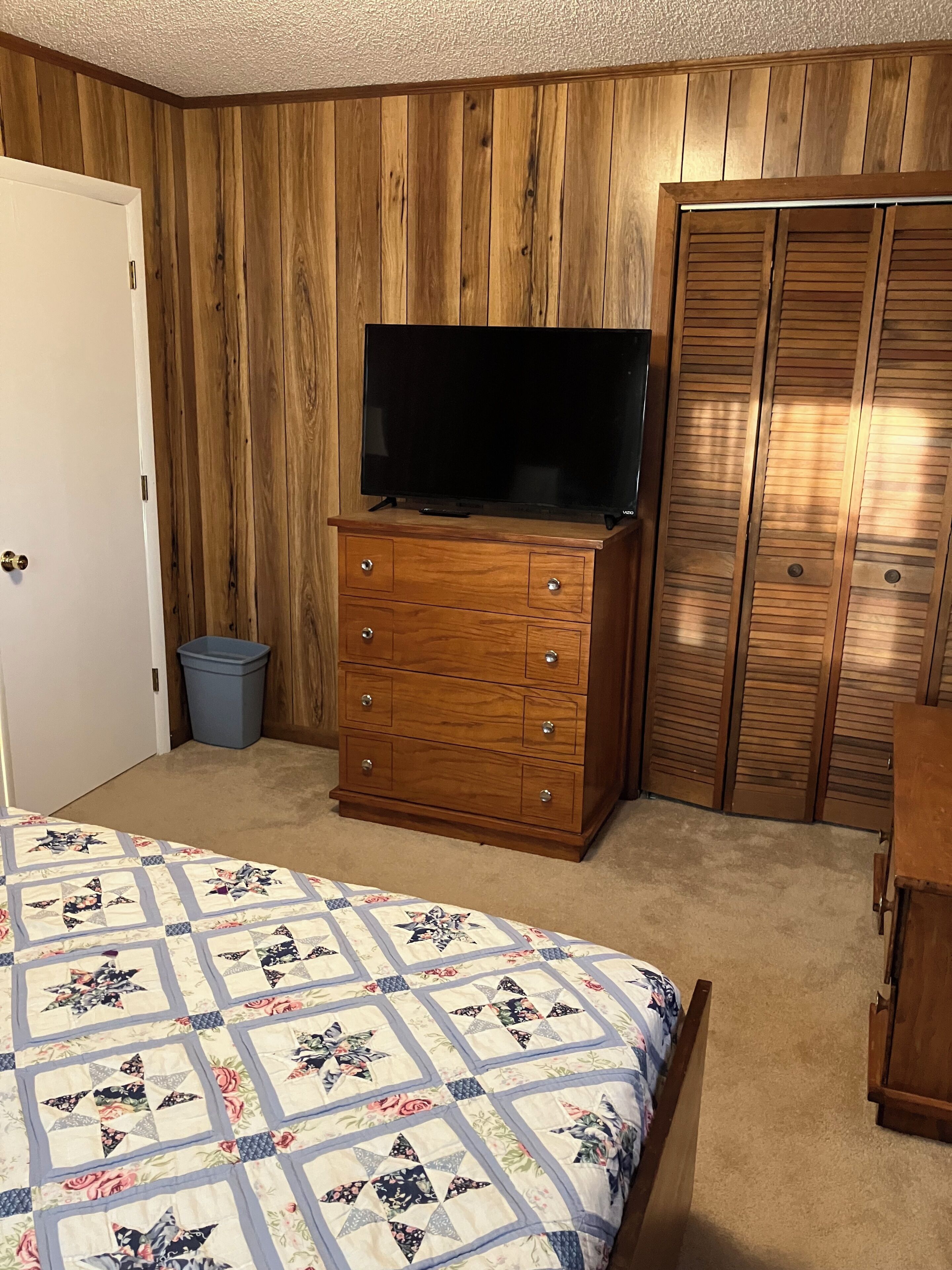 A bedroom with a wooden wardrobe, a television on a dresser, a bed with a floral quilt, and a blue trash bin.
