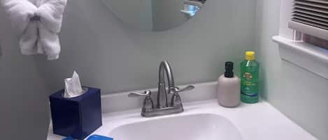 Un baño con un espejo redondo, un lavamanos, un perchero para toallas, una caja de pañuelos y una botella de jabón para manos.