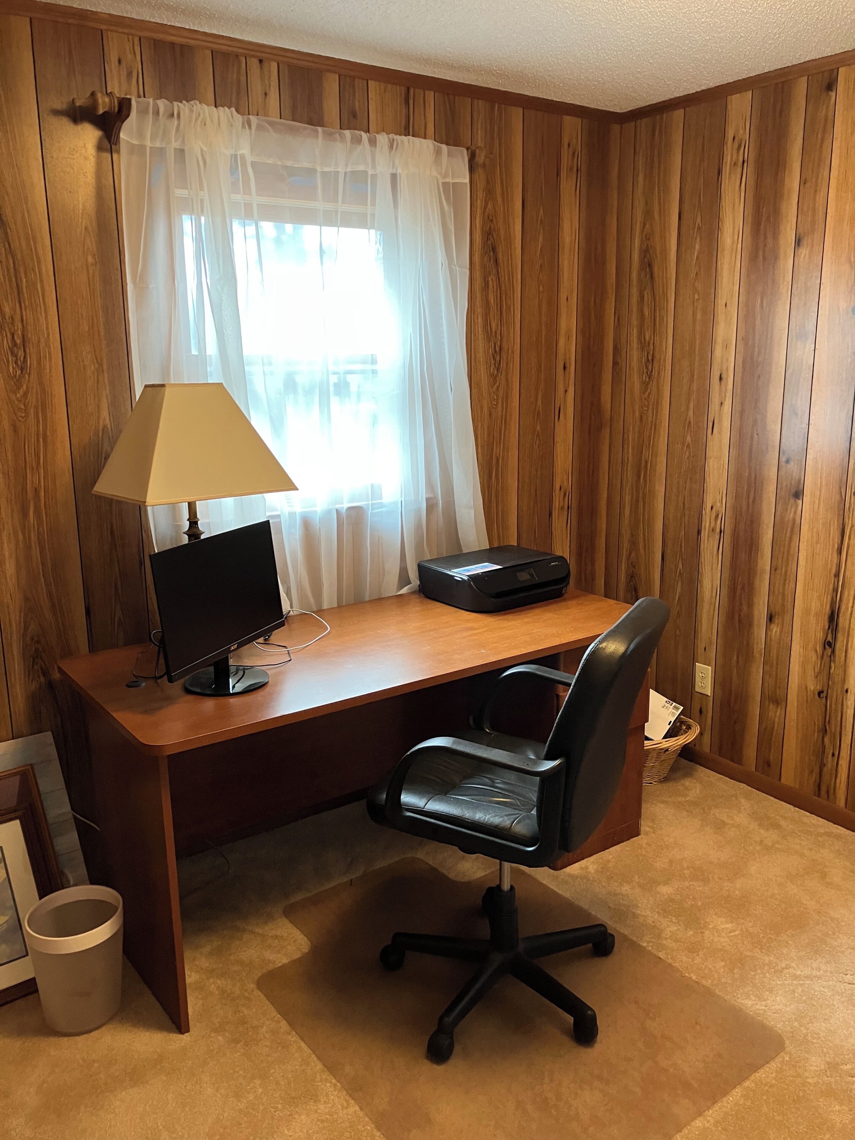 A wooden-paneled room with a desk, a computer monitor, a lamp, and a black office chair.