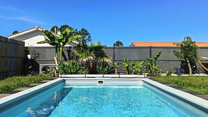 Une piscine rectangulaire à l’eau d’un bleu limpide, entourée d’une clôture en bois, avec une maison et des palmiers en arrière-plan.