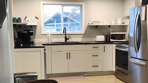 A modern kitchen with white cabinets, a stainless steel refrigerator, a microwave, and a window with a view of a house.