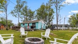 A house with a porch, a fire pit, and three white Adirondack chairs in a grassy yard.