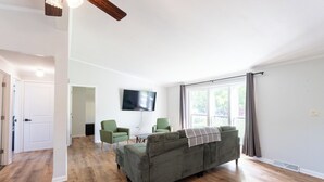 Un salon comprenant un canapé, deux fauteuils, un téléviseur à écran plat et une grande fenêtre avec des rideaux.