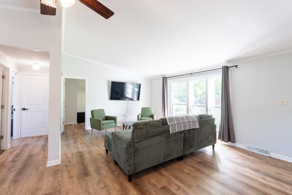 A living room with a sofa, two chairs, a flat-screen TV, and a large window with curtains.
