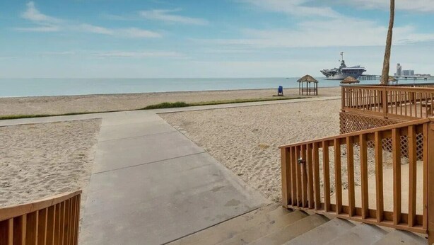 Una playa con un camino de madera, palmeras y un barco a lo lejos.