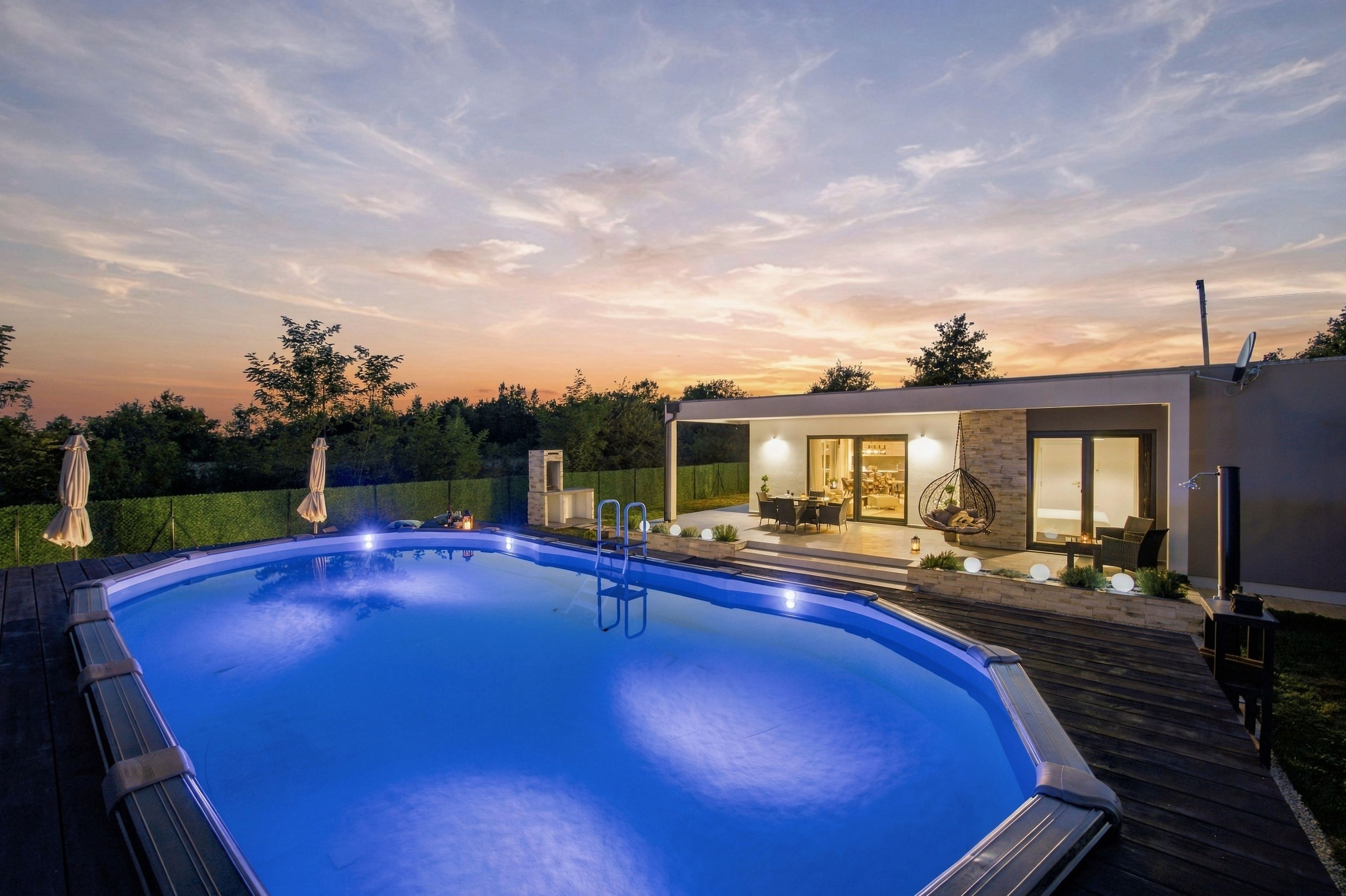 Uma piscina moderna ao ar livre com deck de madeira, um alpendre envidraçado com assentos e um céu ao pôr do sol.