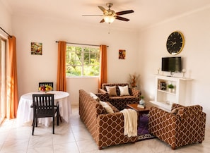 A living room with a sofa set, a dining table with chairs, a ceiling fan, a TV on a shelf, and artwork on the walls. - • “Breathtaking Piton & Ocean Views, Romantic Balcony Retreat (Soufrière)