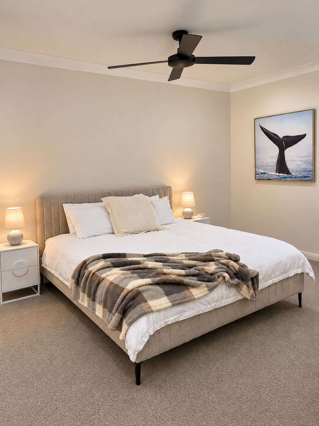 A bedroom with a bed, a ceiling fan, bedside lamps, a nightstand, and a wall-mounted picture of a whale tail.