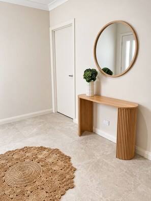 A hallway with a wooden console table, a round mirror, and a woven rug. - SAND - Walk to Marina I Bay Views I 2 Master Suites (Nelson Bay)