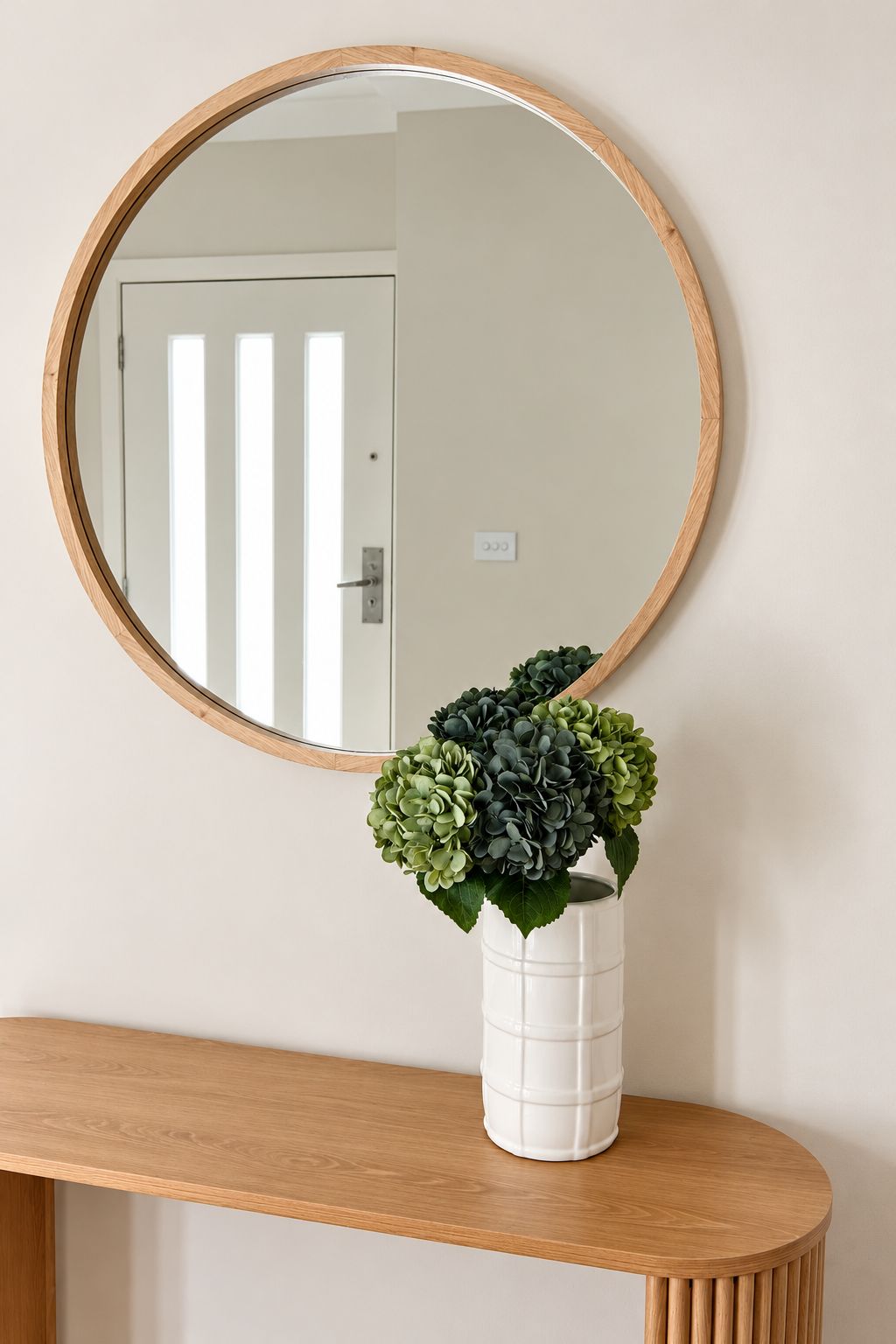 A round mirror with a wooden frame on a wall, a white vase with green foliage on a wooden console table.