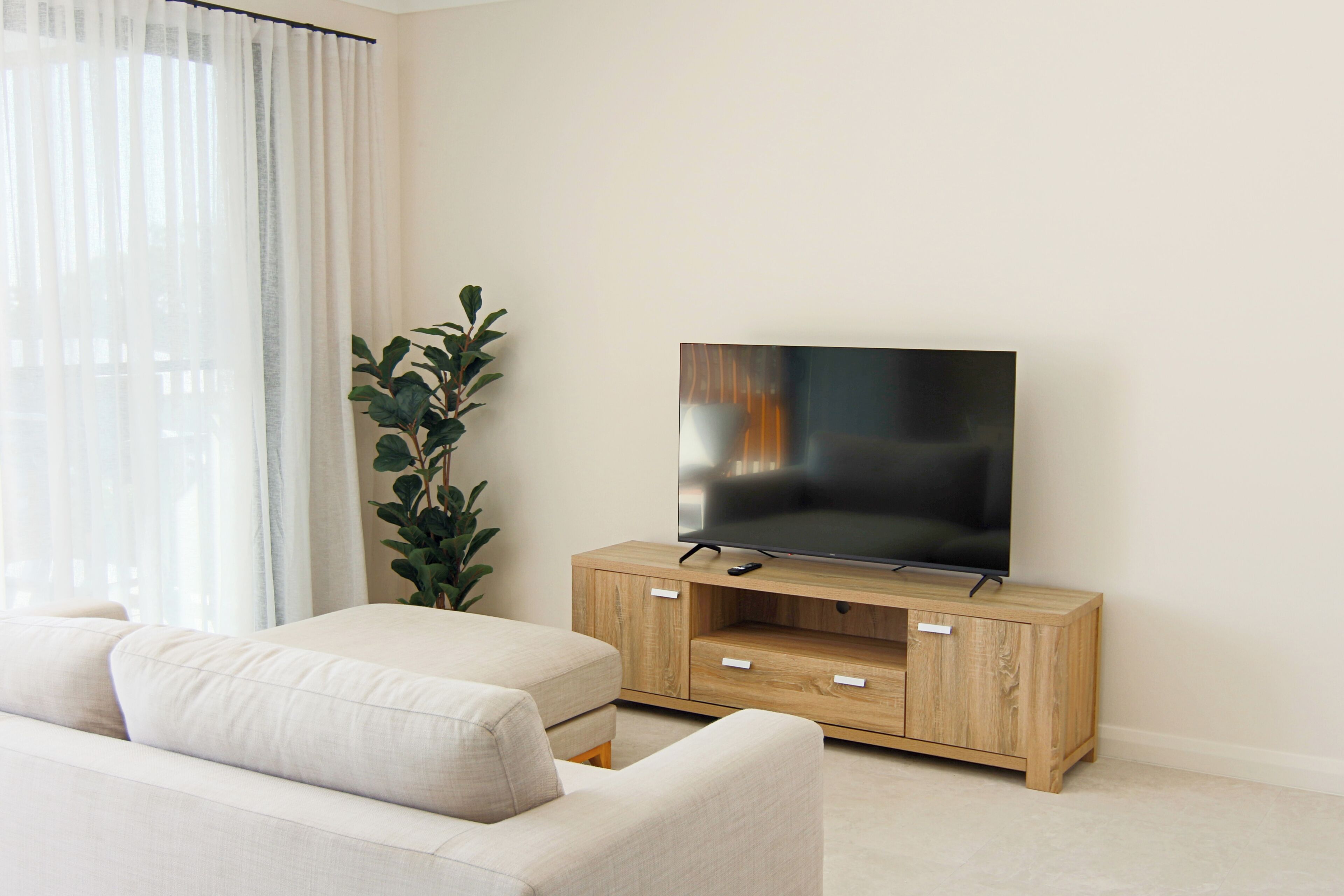 Una sala de estar moderna con un televisor de pantalla plana sobre un mueble de madera, un sofá color beige y una planta en maceta.