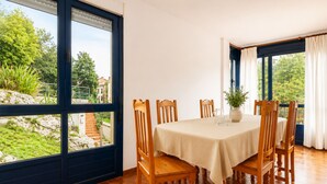 A dining area with a table set for four, large windows with curtains, and a view of greenery outside. - House near Barro Beach Llanes | El Sumidoriu (Barro (Llanes))