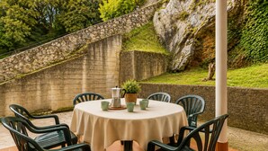A round table with a beige tablecloth, surrounded by six green chairs, set against a stone wall with greenery. - House near Barro Beach Llanes | El Sumidoriu (Barro (Llanes))