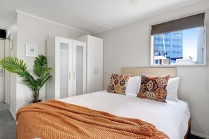Une chambre d’hôtel avec un lit, une armoire blanche, une fenêtre donnant sur un immeuble et une plante en pot.