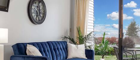 Um sofá azul, uma mesa redonda, um relógio de parede e vista para o exterior através de uma porta de vidro.