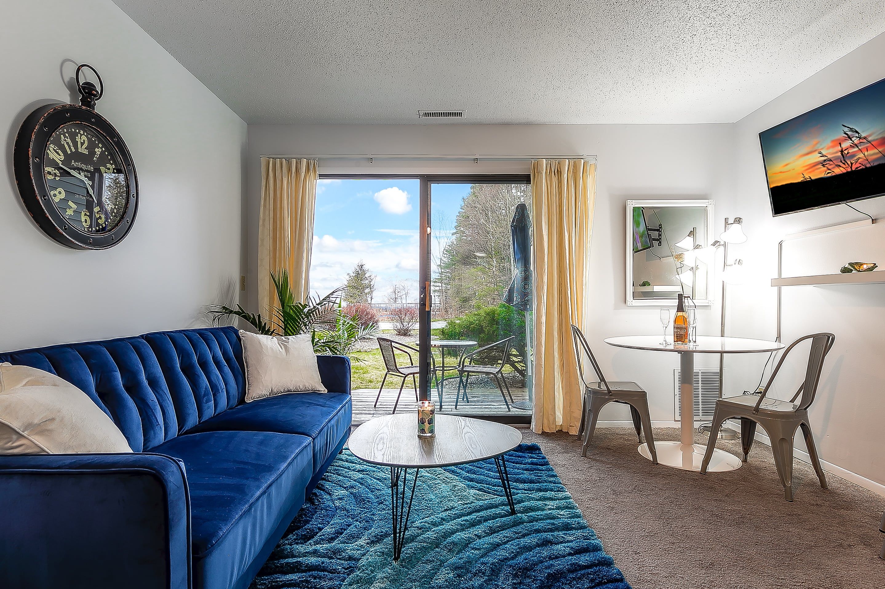 Un salon moderne avec un canapé bleu, une table basse et une vue sur l’extérieur, accessible par une porte vitrée.