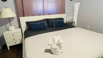 Een netjes opgemaakt bed met een witte deken en kussens, een bedlampje, een nachtkastje met lades en een muur met een decoratieve anker en scheepsschroef.