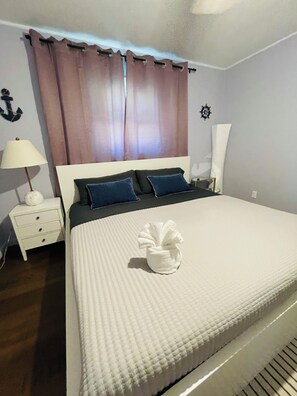 Una cama bien tendida con edredón blanco y almohadas, una lámpara de noche, una mesita de noche con cajones y una pared con un ancla y una rueda de barco decorativos.