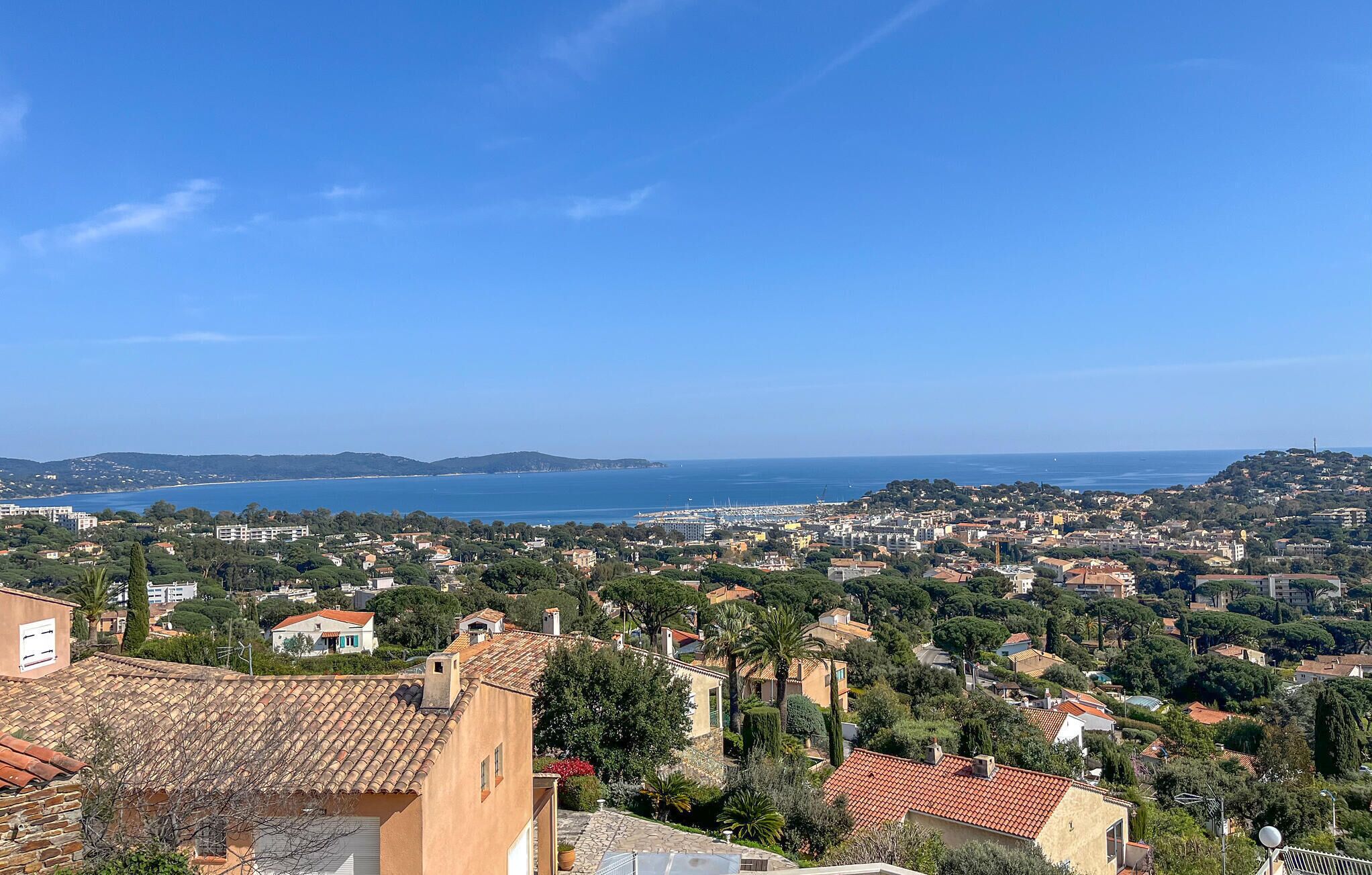 A coastal town with terracotta-roofed buildings, a marina, and a clear blue sky.