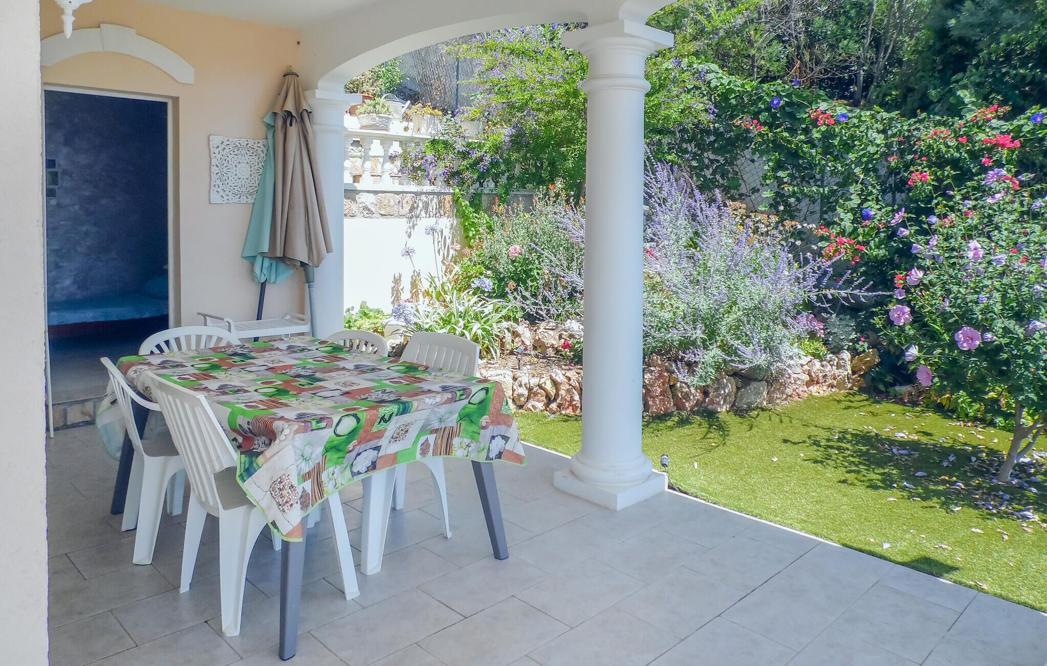 Een overdekt terras met een tafel gedekt voor twee, omringd door witte stoelen, en een tuin met verschillende planten en bloemen.