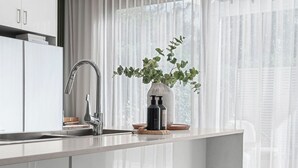 Een moderne keuken met een groot eiland, roestvrijstalen apparatuur en uitzicht op het buitenleven door lichte gordijnen.