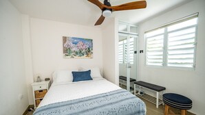 A bedroom with a bed, a nightstand, a bench, a ceiling fan, a window with blinds, and a painting on the wall.