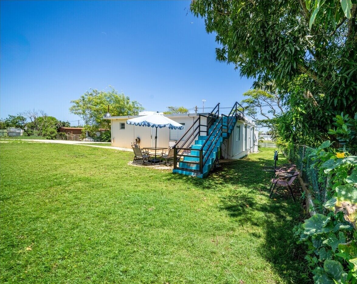 Una casa amb una escala blava que condueix a una zona exterior coberta, un jardí i un petit pat amb cadires.