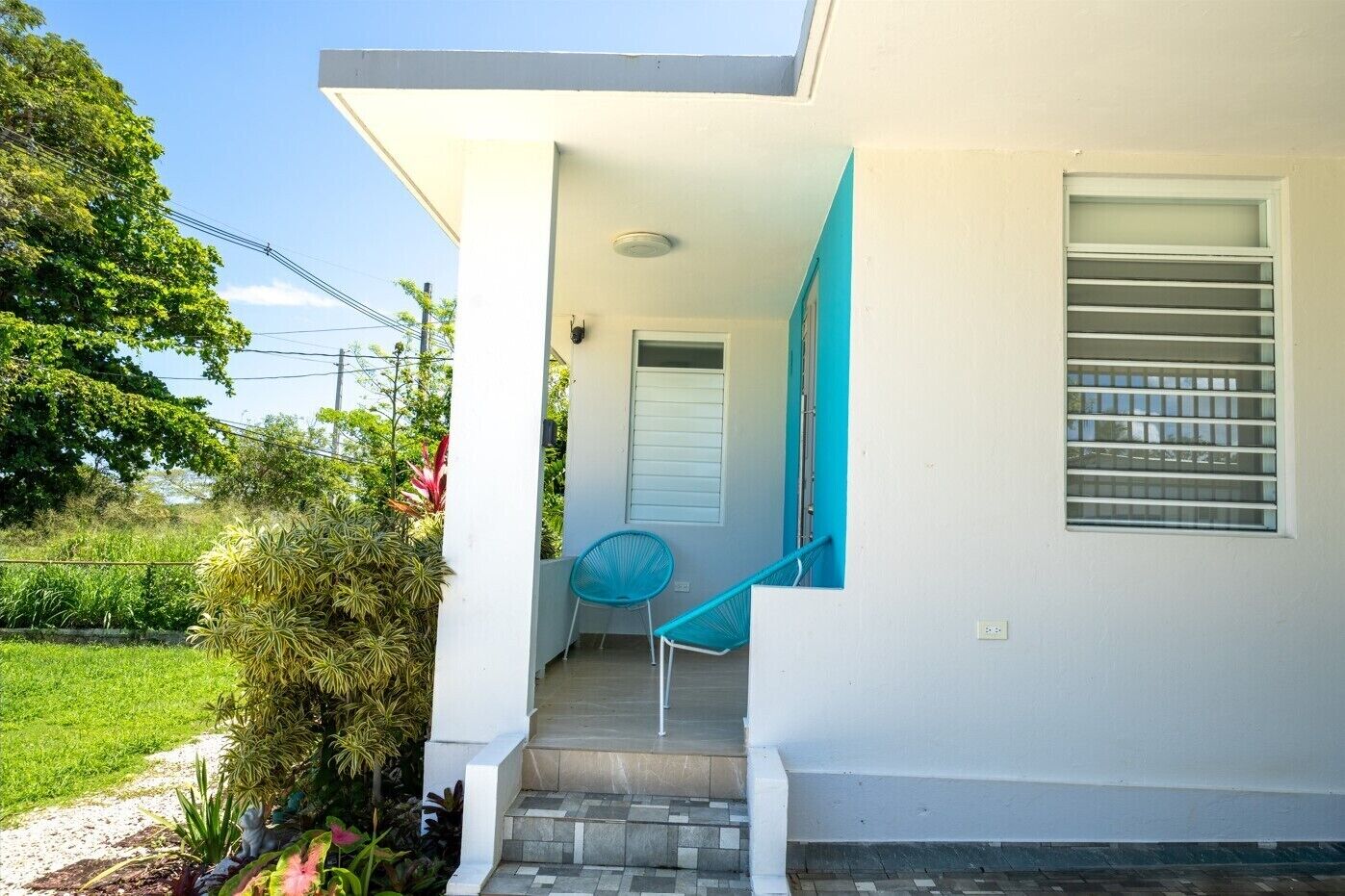 Una casa con una escalera que lleva a una puerta, una ventana con persianas y un pequeño jardín con plantas.