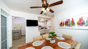 A dining area with a wooden table, four chairs, a ceiling fan, a television, and a colorful cityscape painting on the wall.