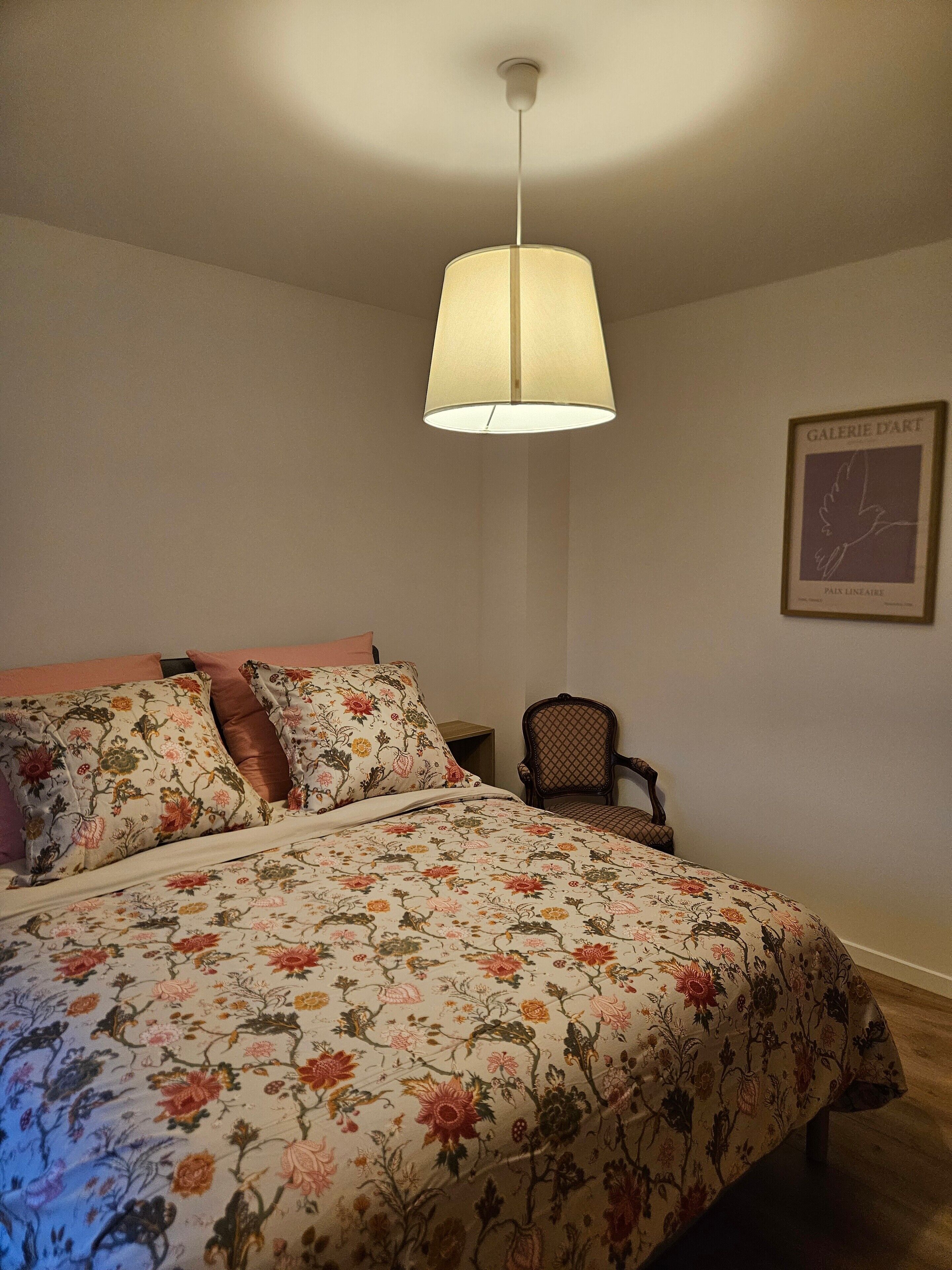 Ein Schlafzimmer mit floralem Bettbezug, einer Hängelampe, einem Stuhl und einem gerahmten Bild an der Wand.