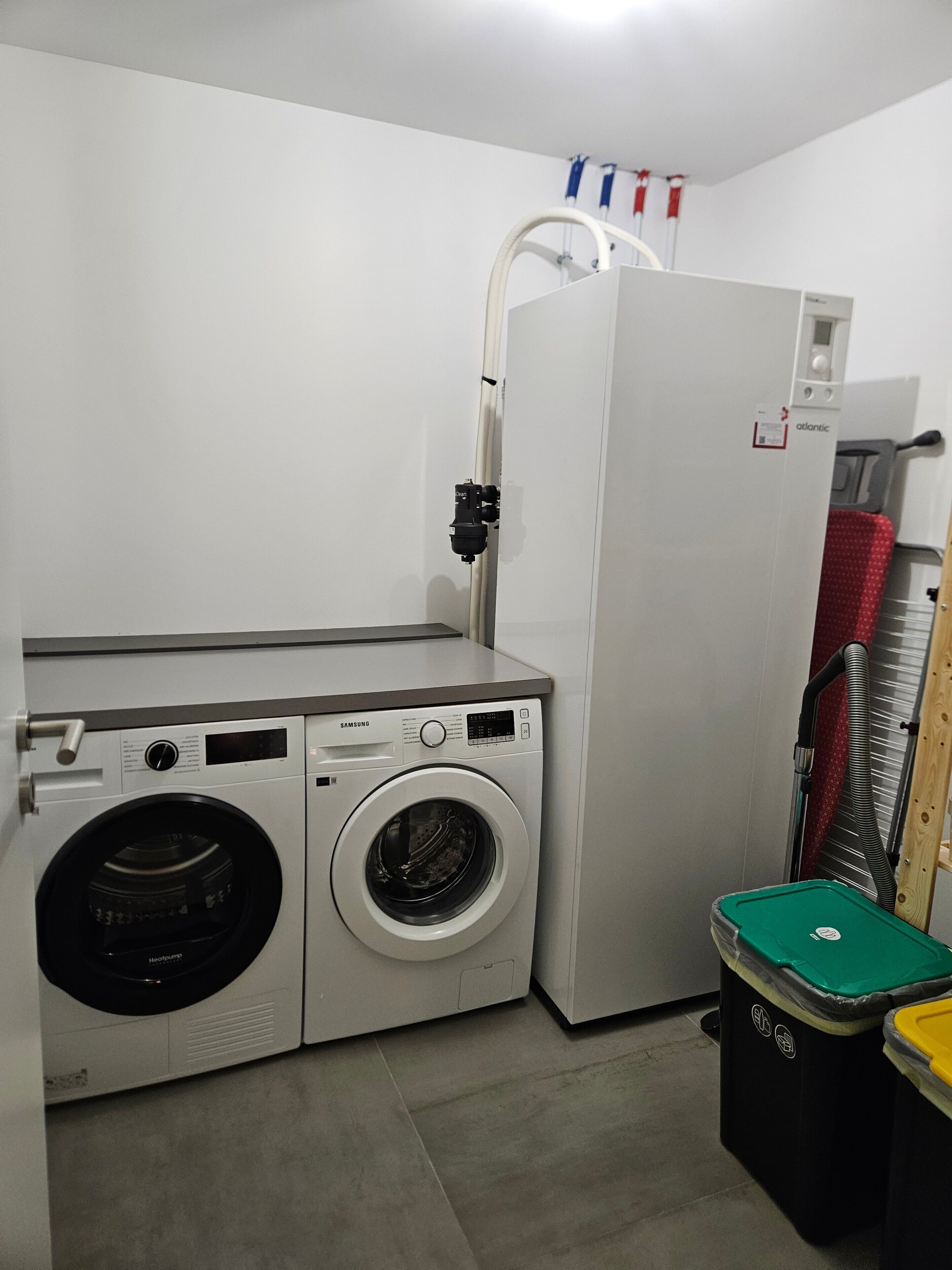 Une petite buanderie équipée d’un lave-linge et d’un sèche-linge Samsung, d’un réfrigérateur et d’une poubelle.