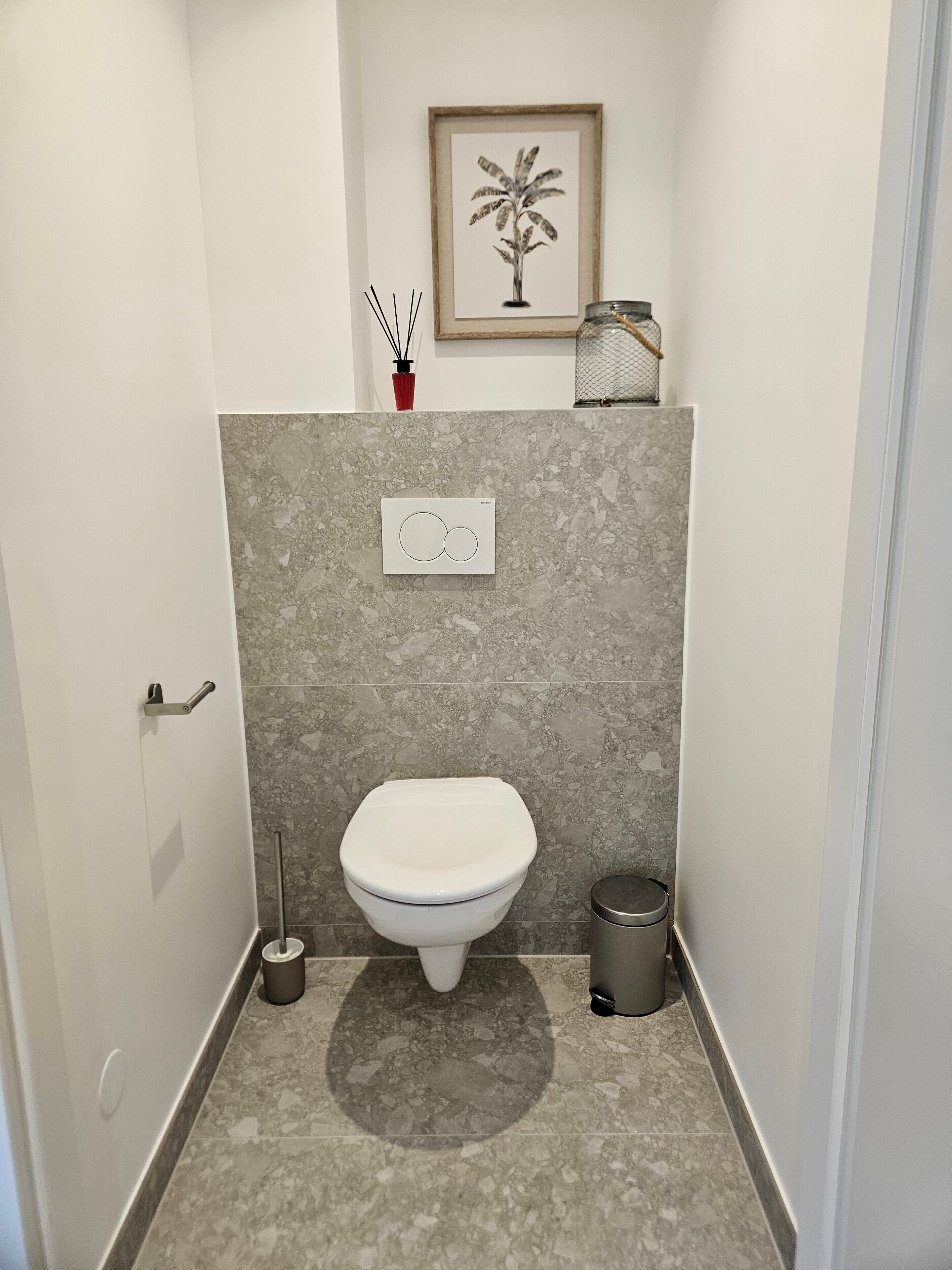 A bathroom with a toilet, a bidet, a trash bin, a framed plant, and a thermostat on the wall.