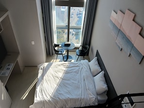 A hotel room with a bed, a small round table, two chairs, a window with curtains, and a view of buildings outside. - Someone Hotel (Yeosu)