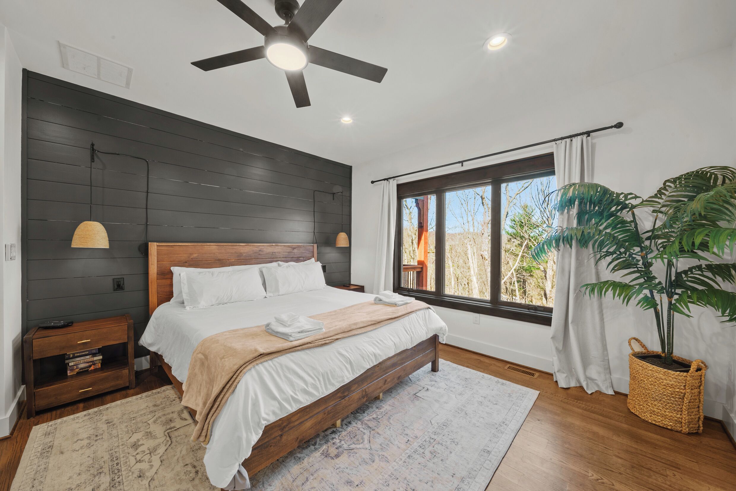 Quarto com cama, mesas de cabeceira, ventoinha no teto, janela com cortinas e um vaso com planta.