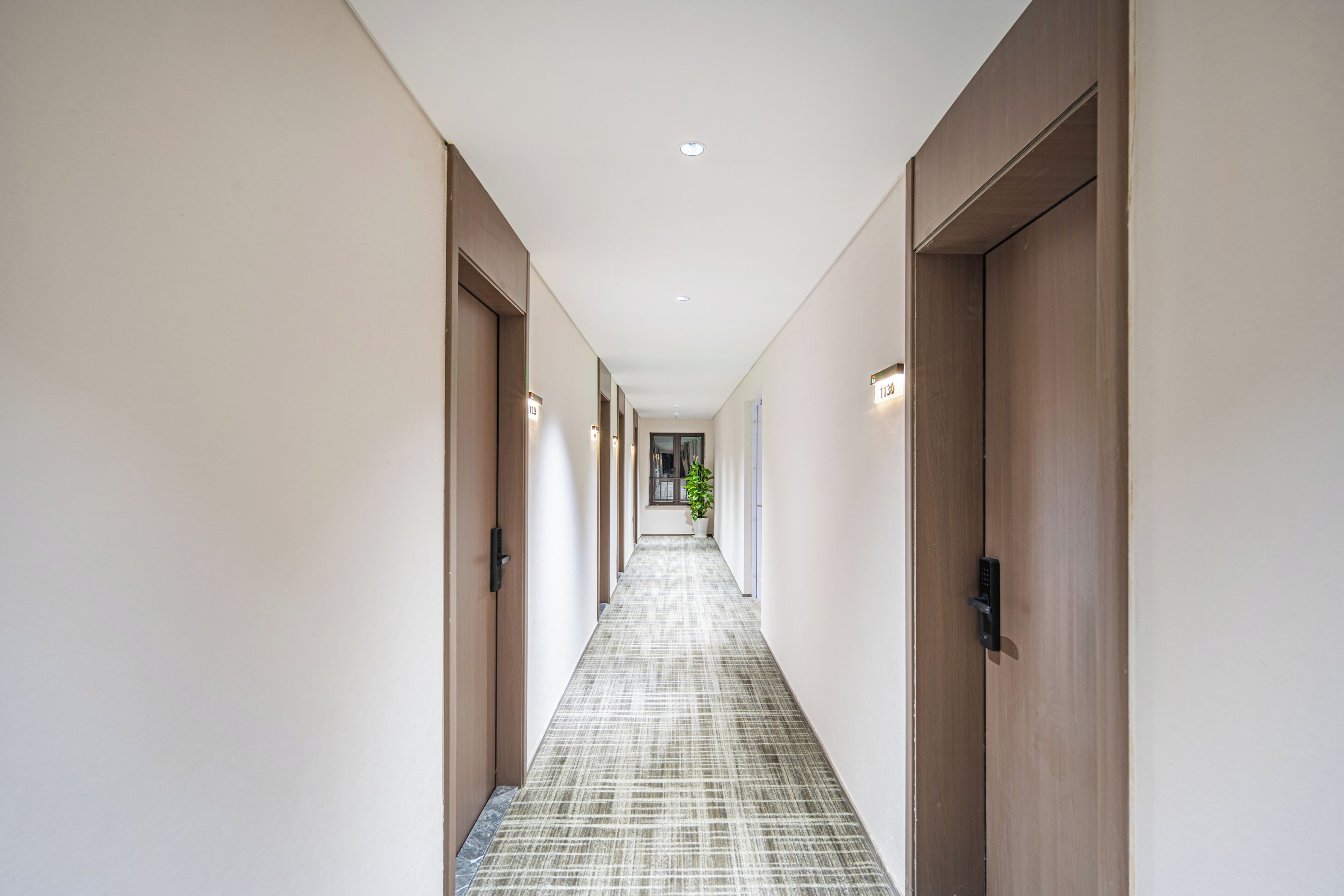 Un pasillo largo y estrecho con puertas de madera a ambos lados, suelo con diseño y luces empotradas en el techo.