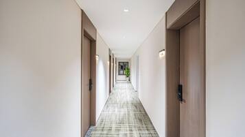 Un long couloir étroit, avec des portes en bois de chaque côté, un sol à motifs et des luminaires encastrés au plafond.