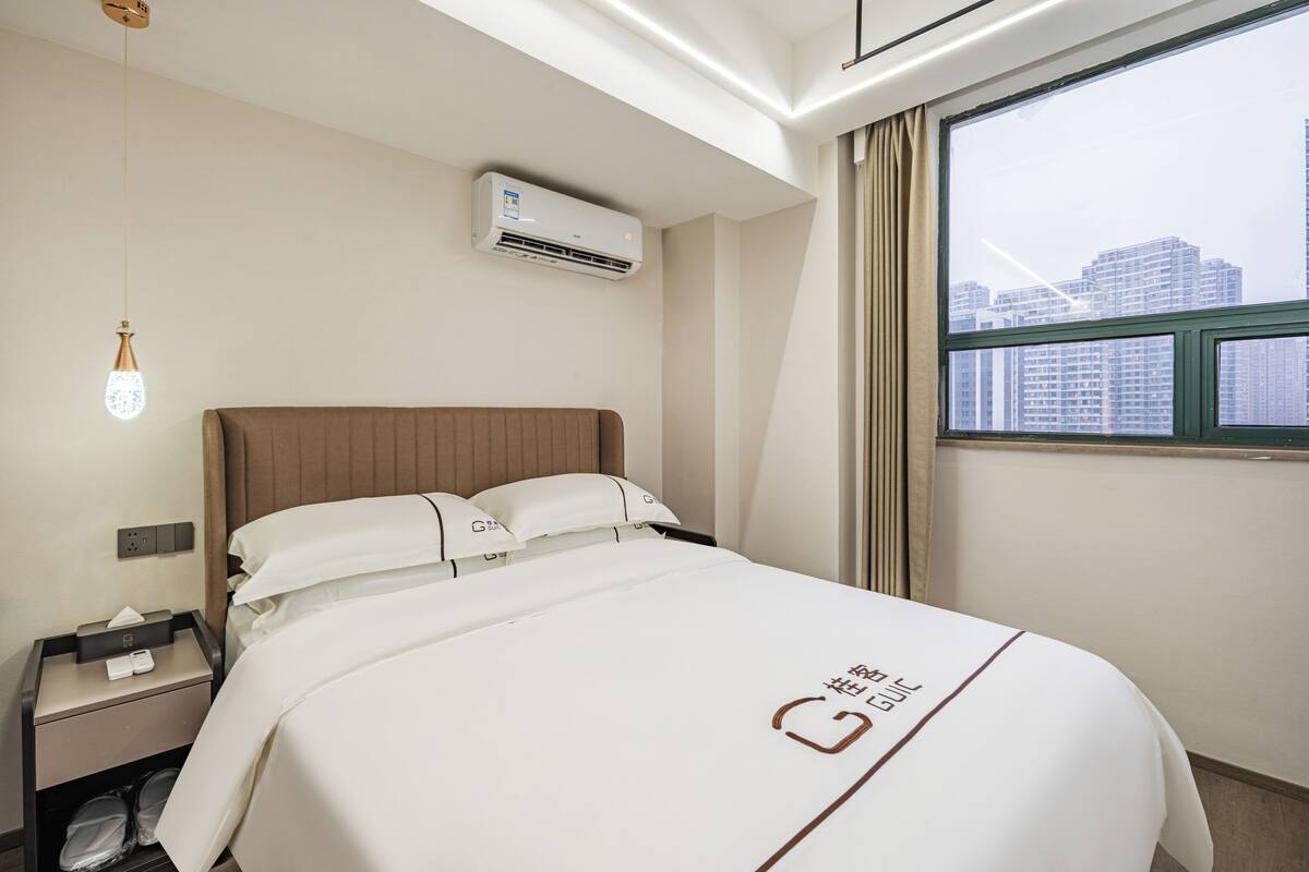 A hotel room with a bed, a nightstand, an air conditioning unit, a window with a city view, and a hanging light fixture.
